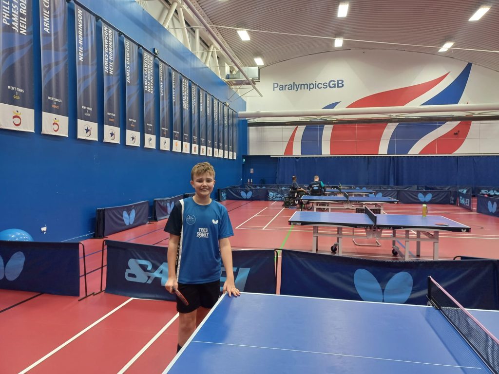 a boy stands by a table tennis table in a top end sports hall featuring a union jack logo emblazoned on the wall behind him