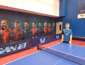 A boy stands by a table tennis table in front of a large picture of elite athletes