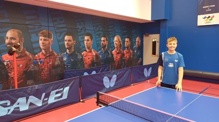 A boy stands by a table tennis table in front of a large picture of elite athletes