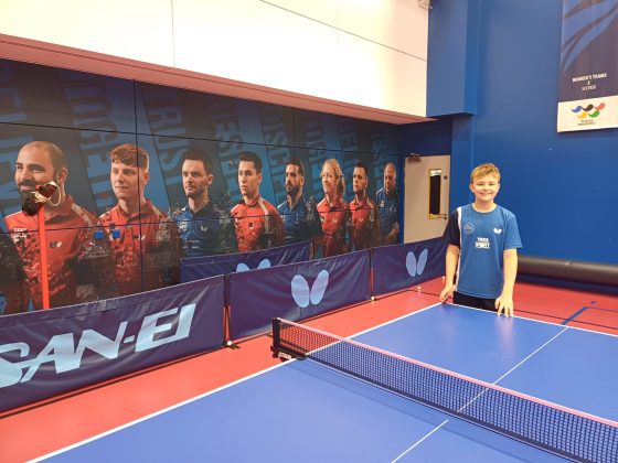 A boy stands by a table tennis table in front of a large picture of elite athletes